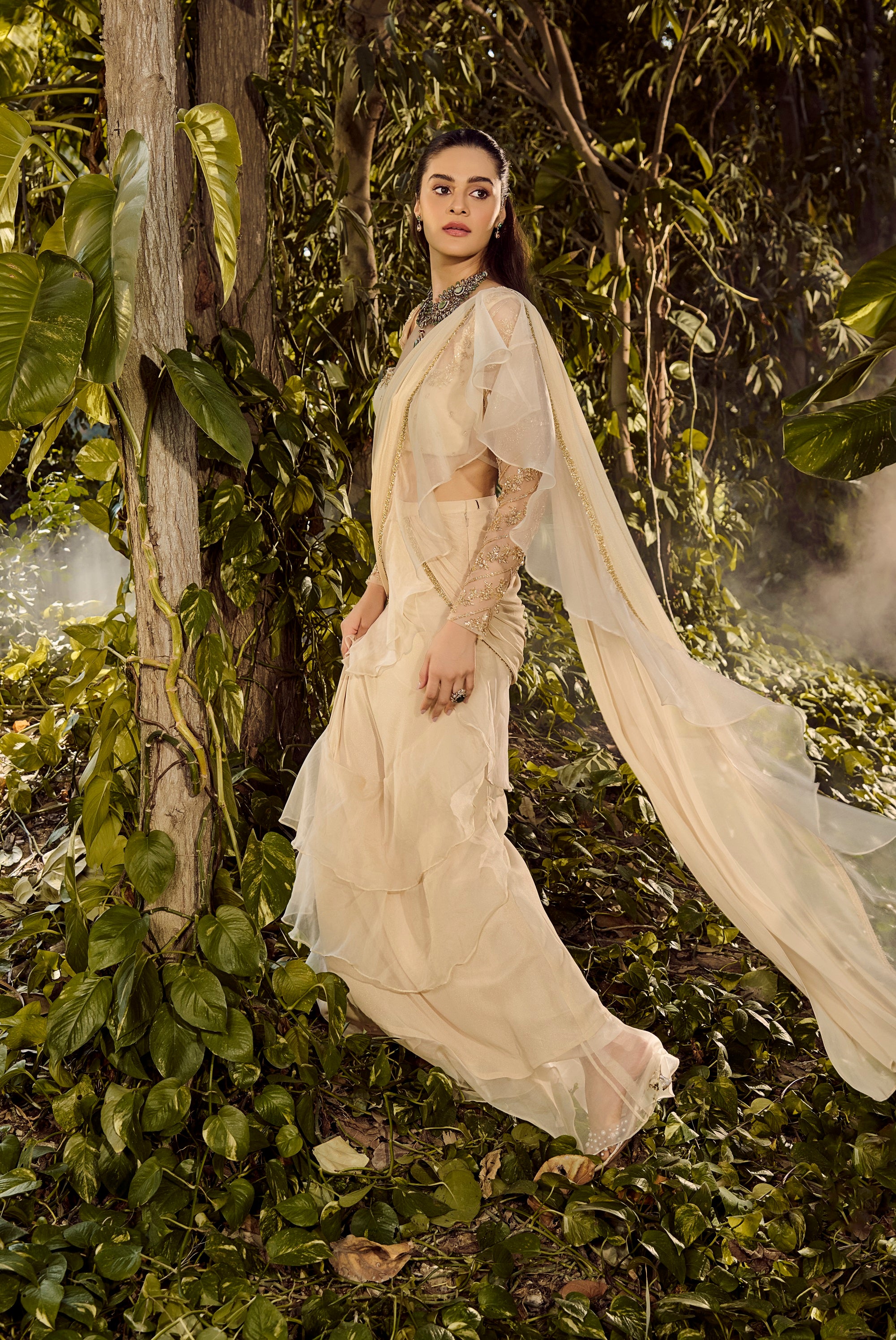 Serene Ruffle Saree. Woman in a flowing white dress standing amidst lush green foliage