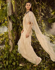 Serene Ruffle Saree. Woman in a flowing white dress standing amidst lush green foliage